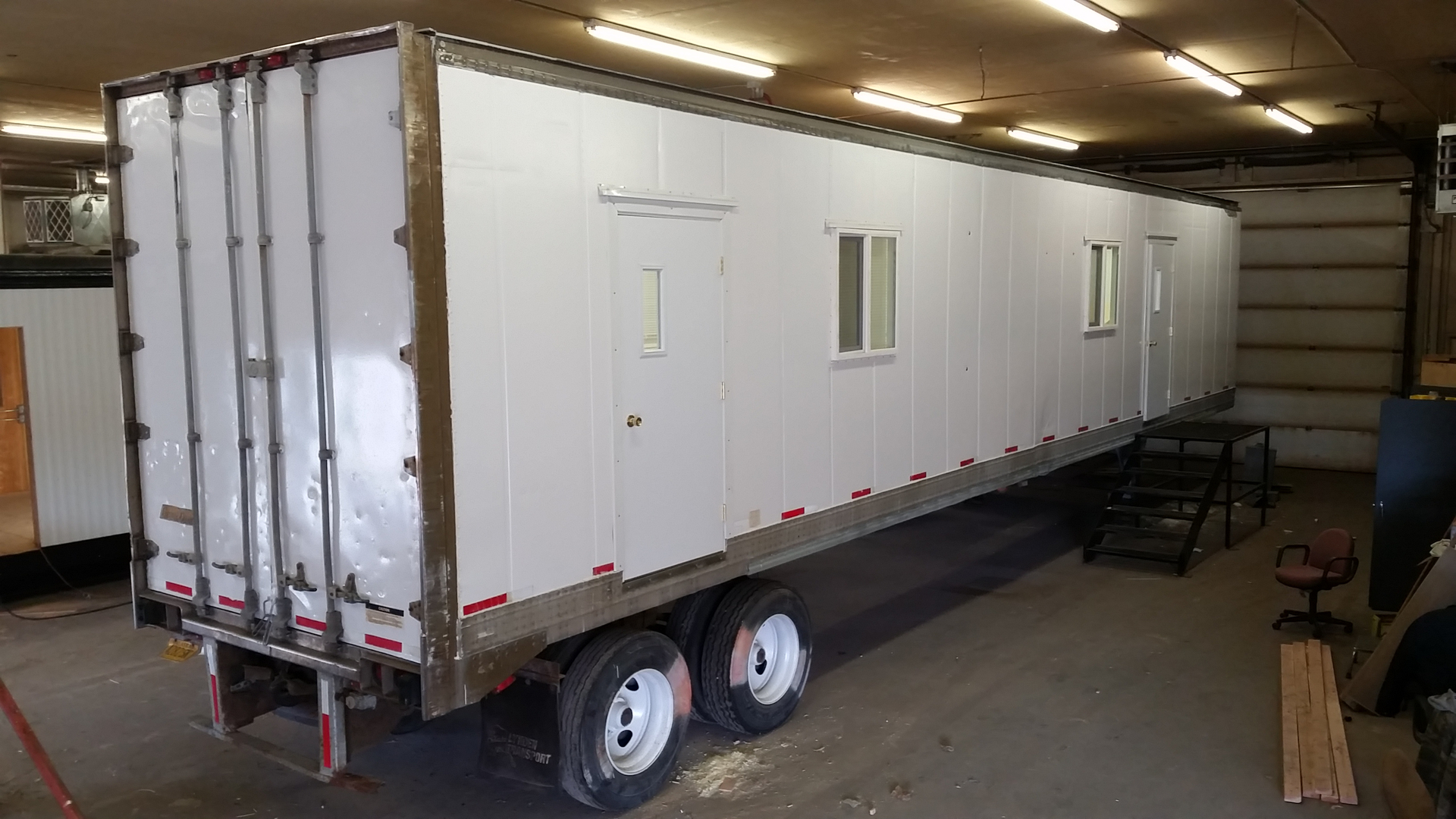 Long white modular office trailer mounted on a semi-trailer inside a warehouse, featuring multiple doors and windows along the side, dual rear axles, and overhead industrial lighting.