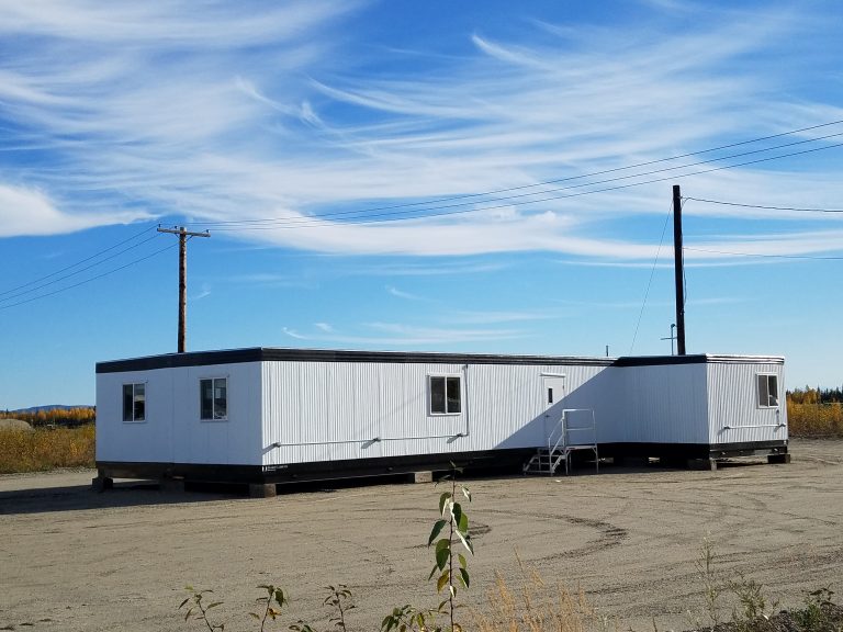 Long modular office building with white metal siding and exterior access steps installed at a remote work site under a clear blue sky