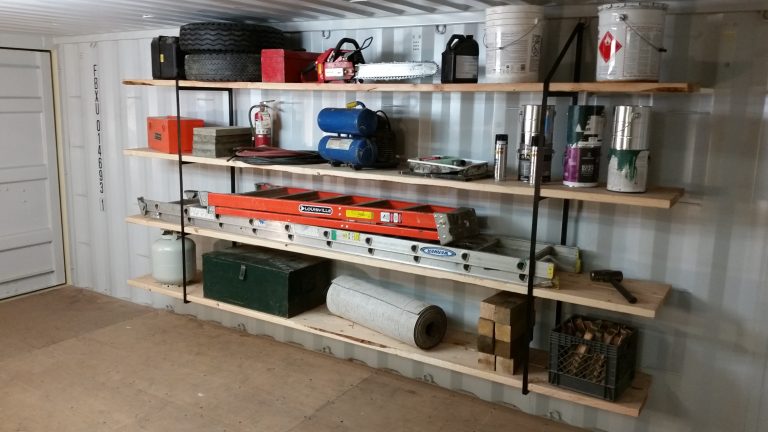 Organized interior of a modified shipping container used as a tool and equipment storage room, featuring wall-mounted shelving with power tools, ladders, paint supplies, and safety equipment arranged for efficient workspace use