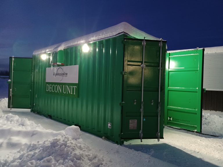Green insulated shipping container labeled “Decon Unit,” illuminated at night in snowy conditions, with exterior lights on and doors open, designed for cold-weather industrial decontamination use.