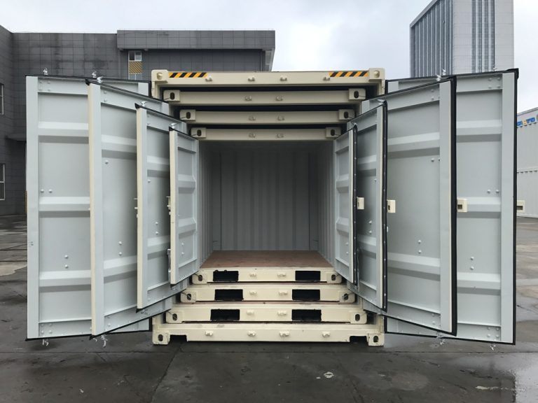 Stacked shipping containers with all front cargo doors open, revealing empty interiors and wooden floors, arranged in an industrial yard with warehouse buildings in the background.