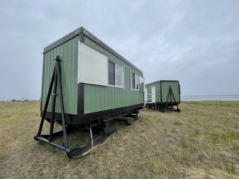 Two green modular skid-mounted buildings with black steel bases sit in an open grassy field, featuring sliding windows and exterior stairs, positioned under an overcast sky in a remote landscape.