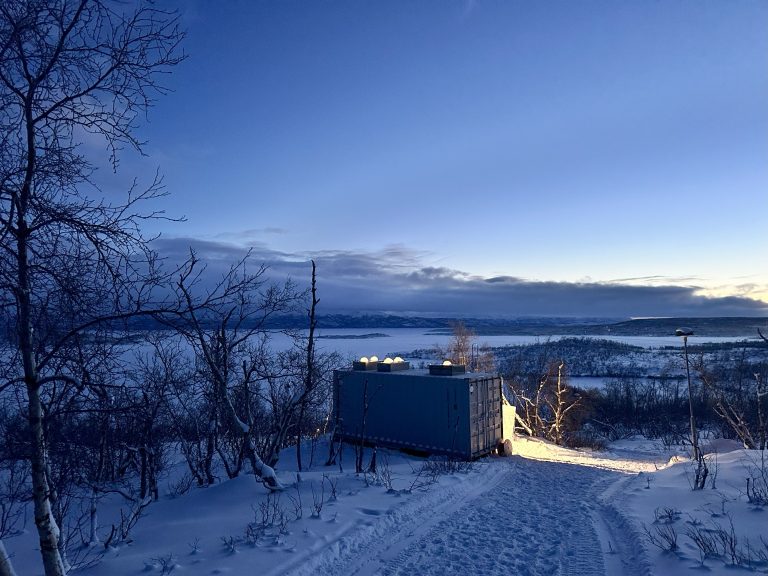 Modular container unit installed in a snowy remote landscape overlooking a frozen lake and mountains, representing off-grid infrastructure in cold climates