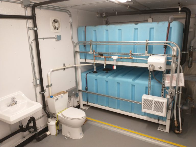 Interior of a modular utility room with integrated water storage tanks, plumbing systems, and bathroom fixtures for off-grid or remote site use